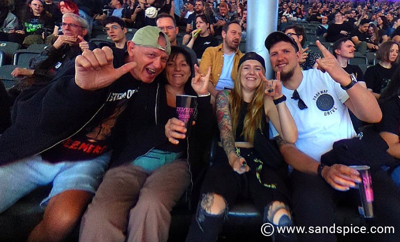 A group of friends smiling and posing for a photo in the tiered seating area at a crowded System of a Down concert in the Berlin Olympiastadion.